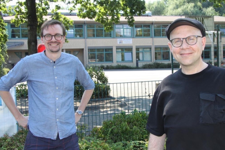 Zwei Männer stehen vor einem Schulhof. Im Hintergrund ist das Gebäude der Grundschule Laukshof zu sehen.