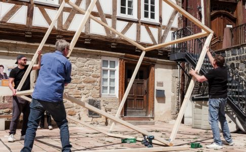 Mehrere Personen bauen einen großen Holzwürfel auf einem Platz vor einem Fachwerkhaus