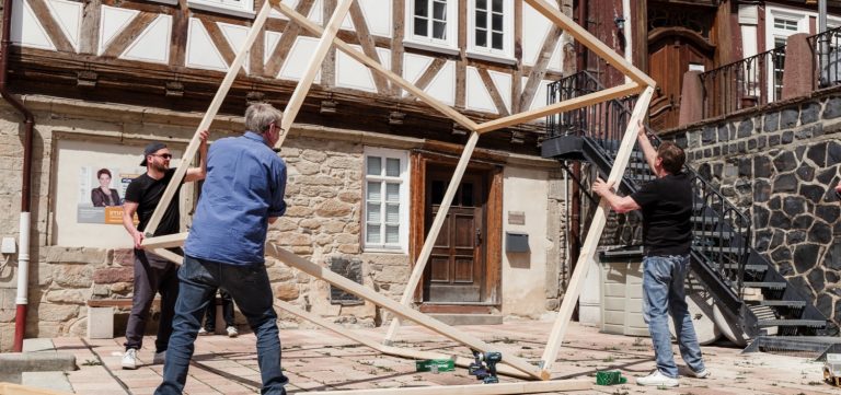 Mehrere Personen bauen einen großen Holzwürfel auf einem Platz vor einem Fachwerkhaus