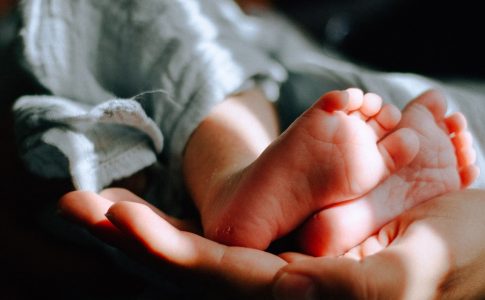 Ein Baby wird von einer erwachsenen Person getragen. Im Vordergrund sind die Babyfüße und die Hand der Erwachsenen zu sehen.