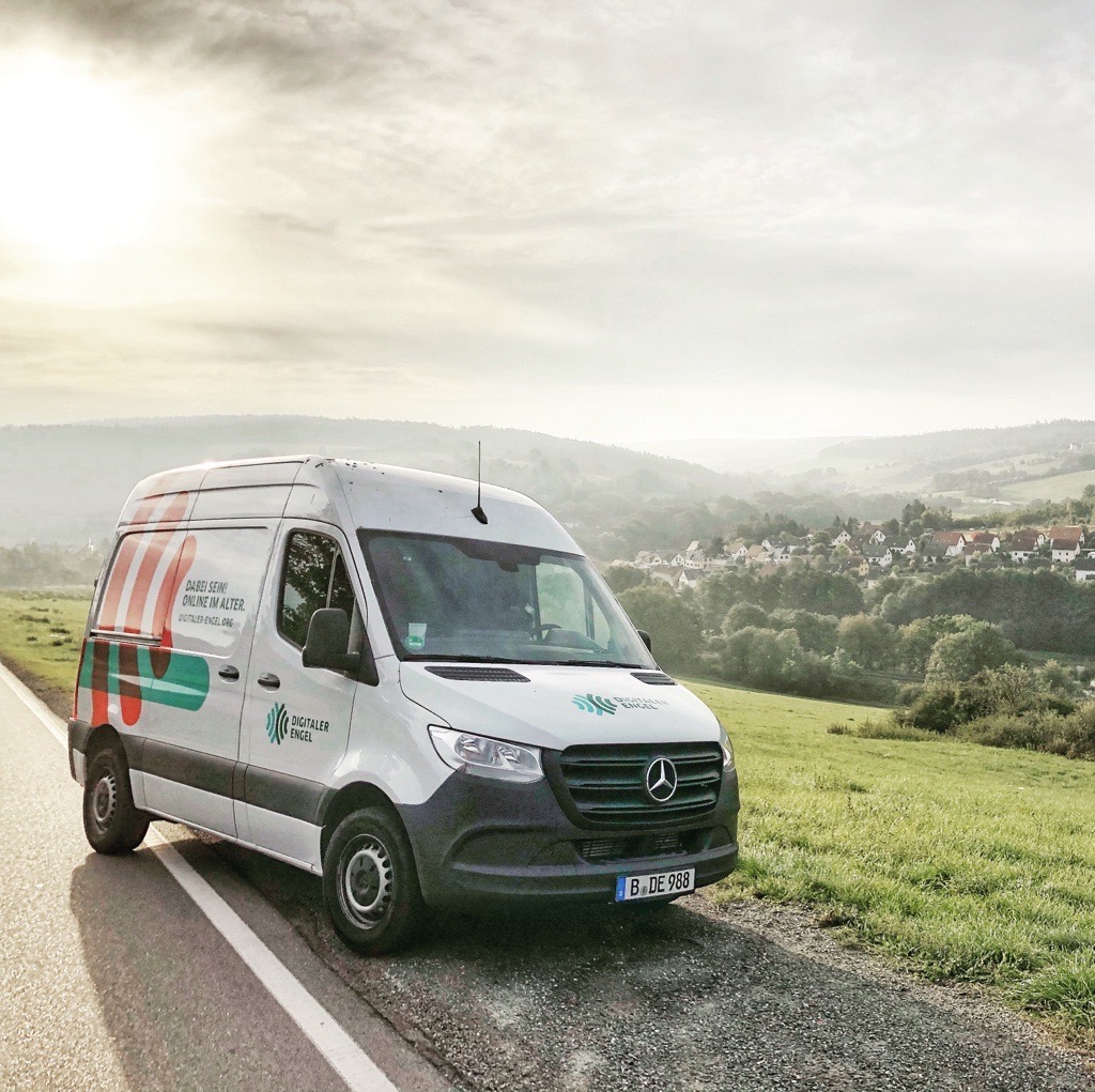 Ein weißer Kleinbus der Organisation Digitaler Engel steht auf dem Standstreifen einer Landstraße. Im Hintergrund ist ein Dorf zu sehen.