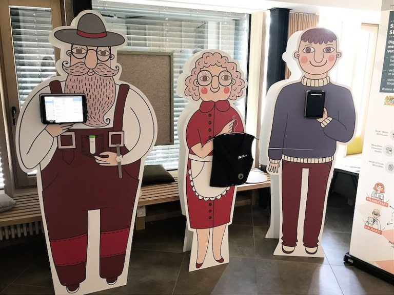 Mit digitalen Displays ausgestattete Pappaufsteller von drei verschiedenen Personen stehen in einem Raum.
