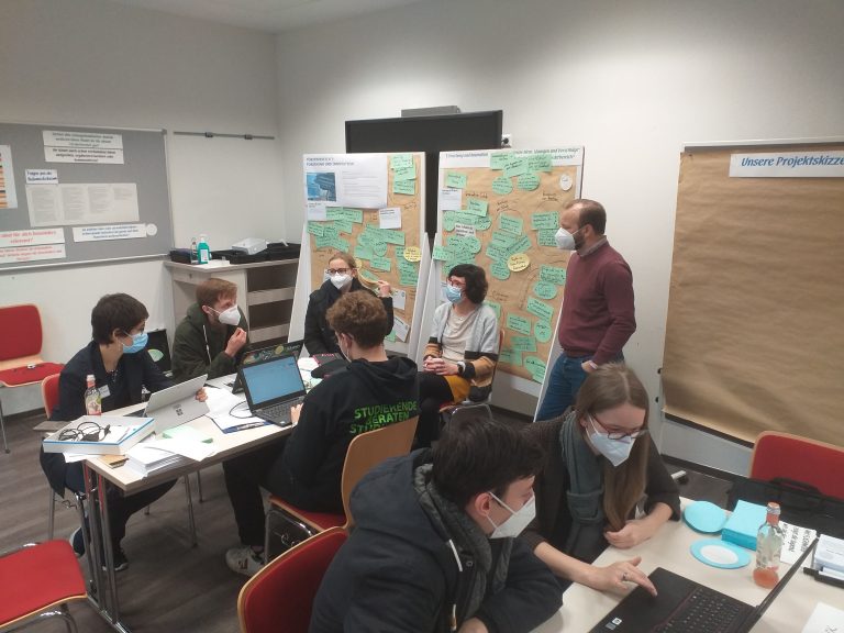 In einem Seminarraum arbeiten junge Menschen an Laptops. Auf einem Board wurden Ideen notiert