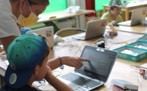 An einem Tisch sitzt ein Junge vor einem Laptop. Eine Frau deutet mit ihrem Finger auf den Bildschirm.