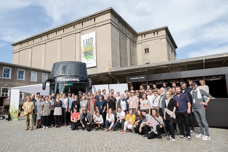 Eine Menschengruppe hat sich vor einem schwarzen Bus positioniert. Im Hintergrund ist ein altes Kühlhaus zu sehen.