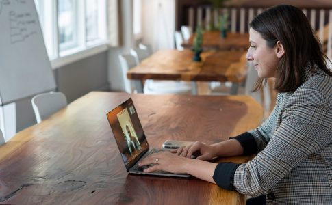 Eine Frau sitzt an einem Tisch und hat einen Laptop vor sich stehen. Auf dem Bildschirm ist eine andere Person zu sehen.