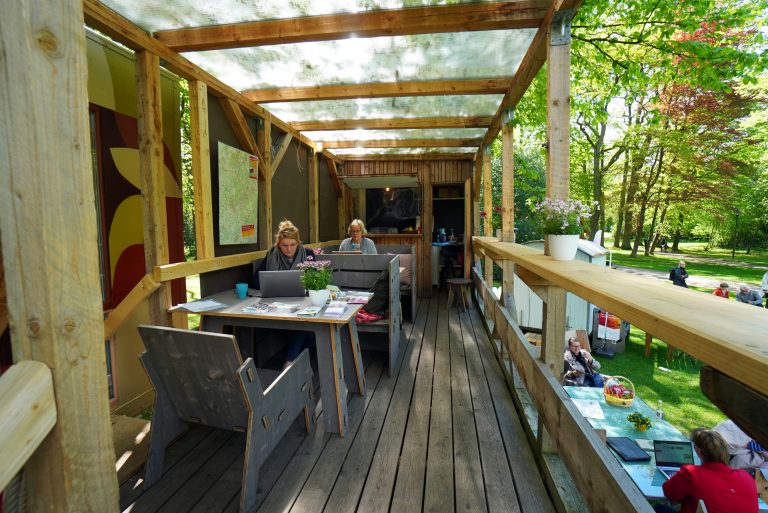 Junge und ältere Menschen sitzen mit Laptops auf einer provisorischen hölzernen Terrasse im Freien.