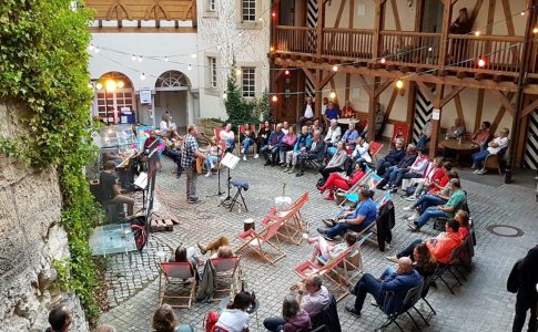 Im Innenhof eines Fachwerkgebäudes sitzen Menschen auf Liegestühlen, vorne steht ein Mann mit Gitarre vor einem Mikrofon.