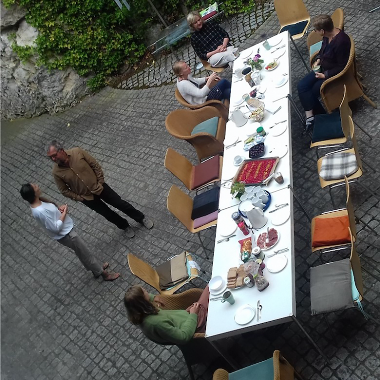 Eine langer, eingedeckter Tisch steht auf Kofsteinpflaster in einem Hof. Auf den bunten Stühlen drumherum sitzen einige Menschen, andere Stühle sind noch frei.