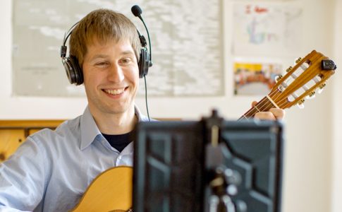 Ein lächelnder junger Mann mit Headset spielt Gitarre und schaut dabei auf ein Tablet.