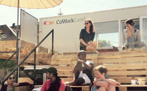 Männer und Frauen sitzen auf Holzpaletten vor einem Container in der Sonne.