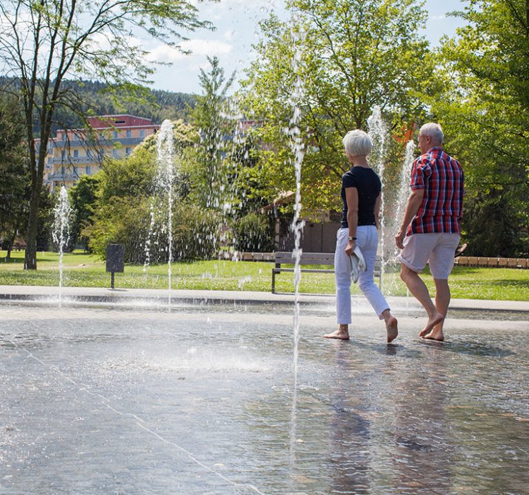 Zwei Menschen laufen barfuß durch einen Springbrunnen in einem grün bewachsenen Park.