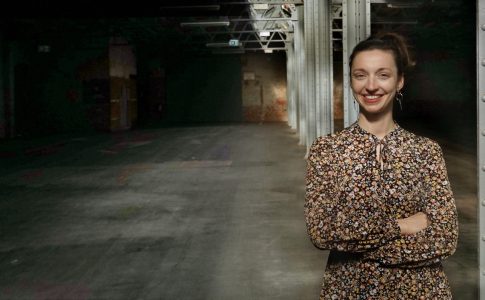 Die Frau Lena Sargalski steht in einer Halle vor einem Stahlträger und lächelt.