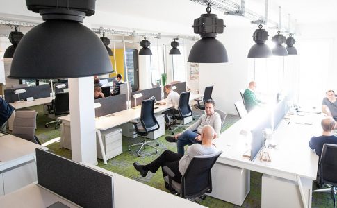 In einem modernen Großraumbüro sitzen Menschen an Computerarbeitsplätzen zusammen und tauschen sich aus.