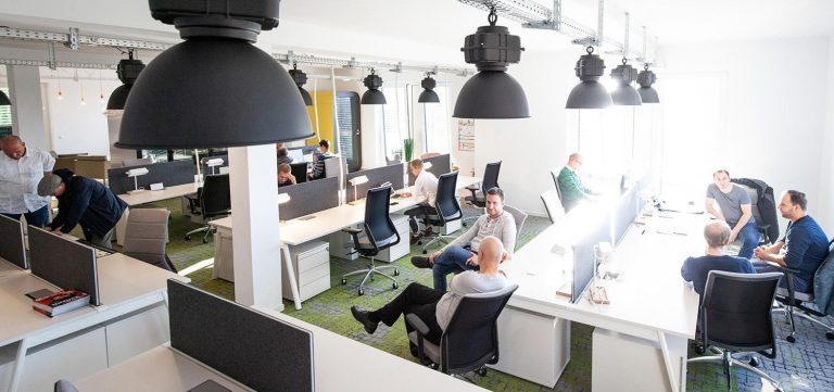 In einem modernen Großraumbüro sitzen Menschen an Computerarbeitsplätzen zusammen und tauschen sich aus.