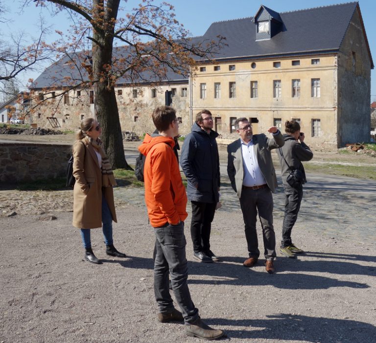 Vor zwei älteren Gebäuden mit brüchiger Fassade steht eine kleine Menschengruppe auf einer Kiesfläche.