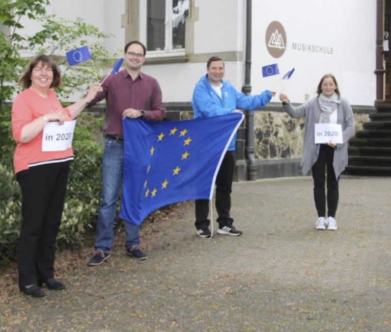 Vier Personen stehen vor einem weißen Gebäude und halten eine große Europafahne.