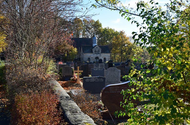 Zu sehen ist ein Friedhof mit Grabsteinen, der von einer Mauer eingefasst ist. Im Hintergrund steht eine Friedhofskapelle.