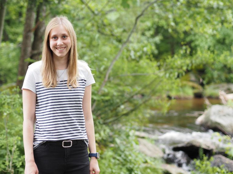 Eine Frau mit langen blonden Haaren steht an einem Bächlein im Wald.