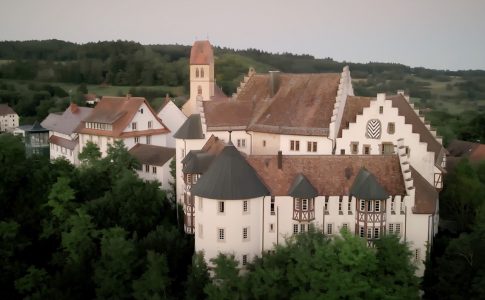 Ein Schloss liegt auf einem Gipfel inmitten grüner Bäume und Hügel.