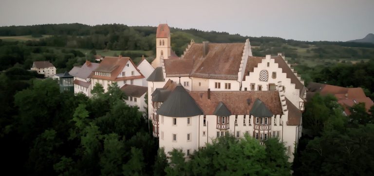 Ein Schloss liegt auf einem Gipfel inmitten grüner Bäume und Hügel.