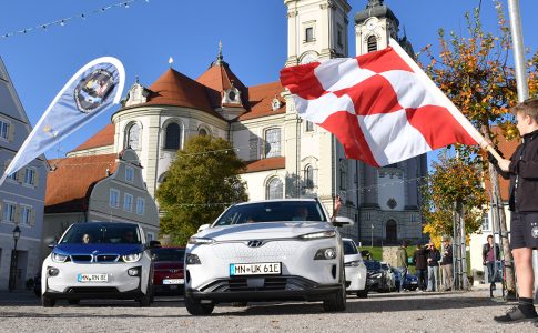 Ein Auto fährt durch eine Kleinstadt, am Rand stehen Menschen mit Fahnen.