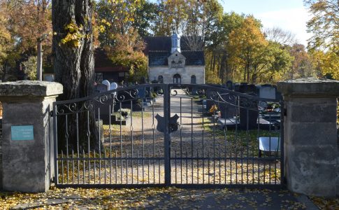 Hinter einem schmiedeeisernen Tor liegt ein Friedhof mit Grabsteinen und einer Kapelle.