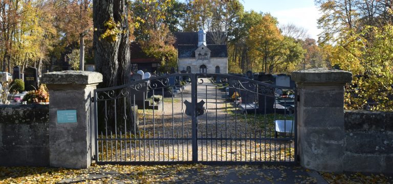 Hinter einem schmiedeeisernen Tor liegt ein Friedhof mit Grabsteinen und einer Kapelle.