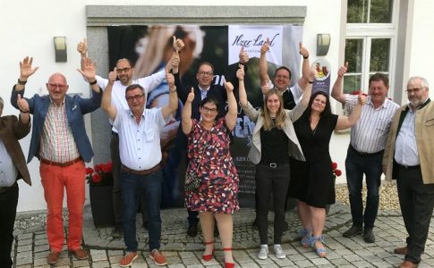 Eine Gruppe Menschen – acht Männer und drei Frauen – steht vor einem weißen Gebäude. Alle recken die Hände in die Höhe.