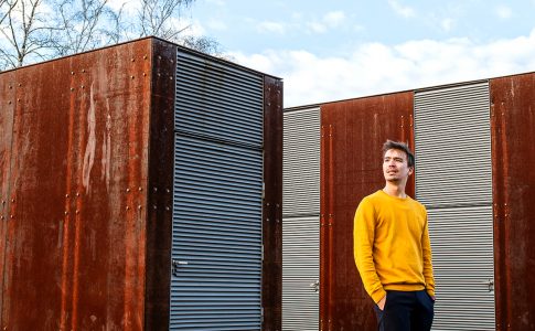 Der Oberstudienrat Bob Blume steht mit einem gelben Sweatshirt auf einer Freifläche zwischen Containern.