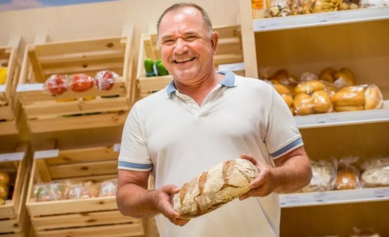 Ein Mann hält ein Brot in der Hand und steht vor einem Backwarenregal.