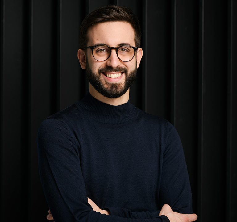 Ein Mann im blauen Pulli lächelt. Er trägt eine dunkle Brille und hat einen schwarzen Vollbart.