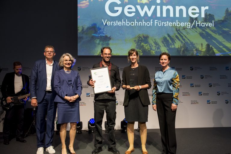 Vor einer bunten Rückwand stehen fünf Personen auf einer Bühne. Ein schwarz gekleideter Mann hält eine Urkunde.