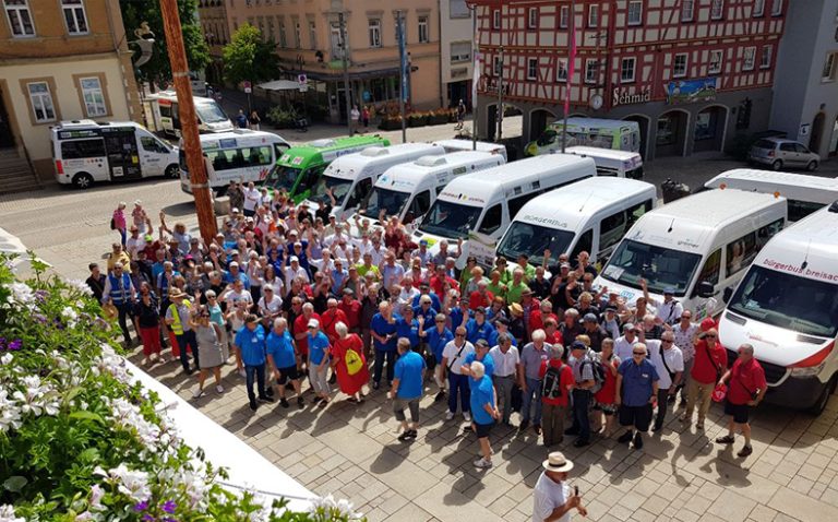 Auf einem Platz stehen weiße Kleinbusse Seite an Seite. Davor eine Gruppe Menschen in bunten T-Shirts.