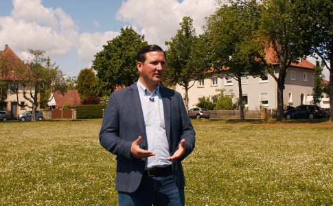 Benedikt Krams steht auf einer Wiese und gestikuliert. Im Hintergrund stehen Wohnhäuser und Bäume.