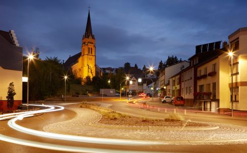 Im Vordergrund ist ein Verkehrskreisel zu sehen, im Hintergrund eine Kirche, dazwischen Wohnhäuser.