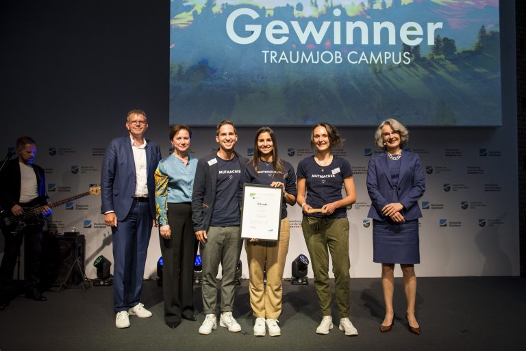 Gewinner eines Wettbewerbs stehen mit Urkunde auf einer Bühne, im Hintergrund eine Leinwand