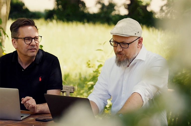 Zwei Männer mit Laptops sitzen vor grüner Kulisse.