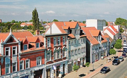 Autos fahren auf einer sonnigen Straße, die von bunten dreistöckigen Häusern mit roten Dächern gesäumt ist.