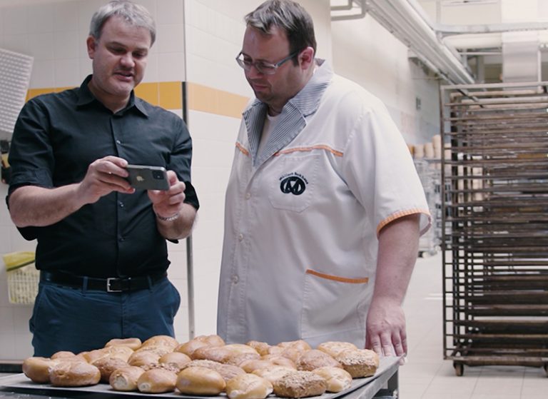 Zwei Männer schauen auf ein Smartphonedisplay. Auf einem Backblech liegen Brötchen.