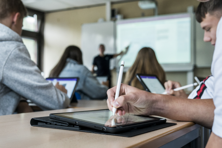 Schülerhand, die mit einem Stift auf ein Tablet schreibt, im Hintergrund zeigt ein Lehrer auf eine digitale Tafel.