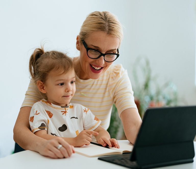 Mutter sitzt mit Kind auf dem Schoß vor einem aufgeklappten Laptop.