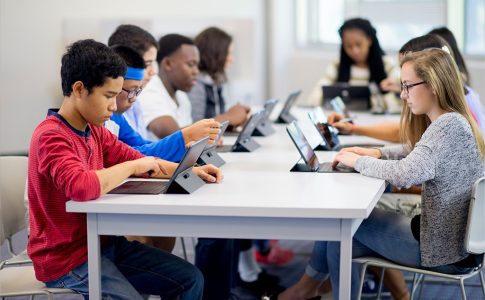 Mehrere Kinder, die in einem Klassenzimmer zusammen um einen Tisch sitzen und an Tablets arbeiten.