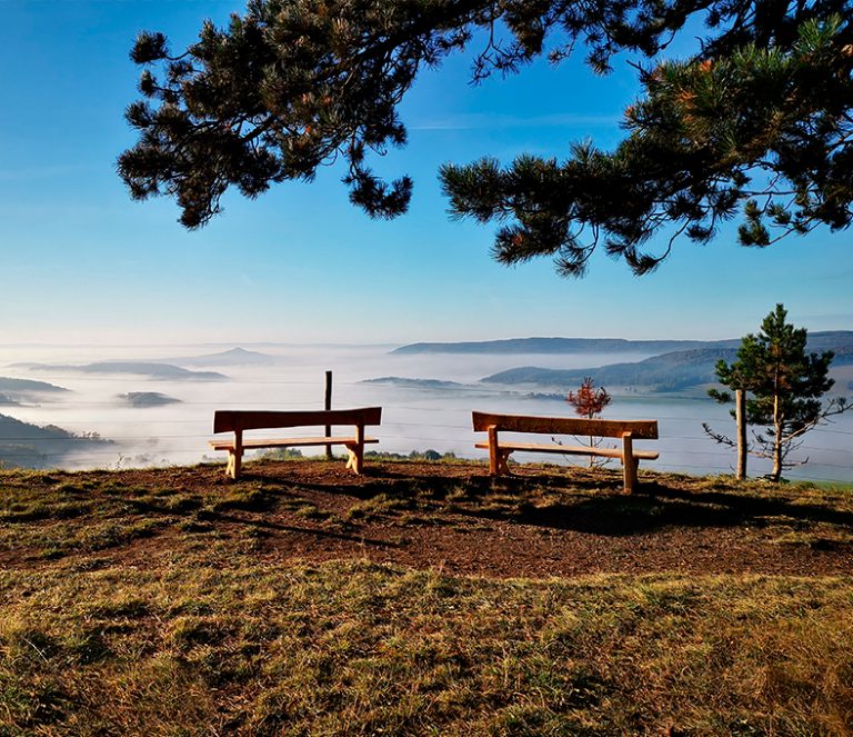 Zwei Bänke auf einer Anhöhe mit Blick auf eine Hügellandschaft, in der die Wolken hängen. Darüber blauer Himmel.