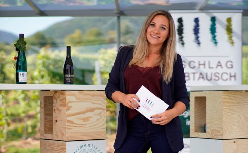 Blonde Frau steht mit Moderationskarten in der Hand vor Weinflaschen und grüner Natur