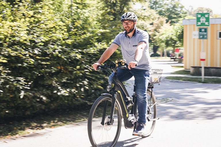 Ein Mann fährt auf einem Fahrrad auf der Straße. Im Hintergrund ist eine Hecke zu sehen.