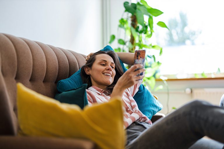 Eine Frau sitzt auf dem Sofa und schaut lachend in ein Smartphone.