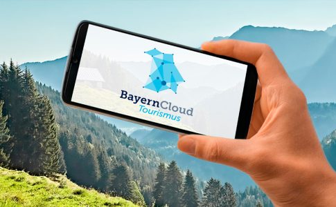 Hand, die ein Smartphone mit dem Logo der BayernCloud vor ein Bergpanorama hält