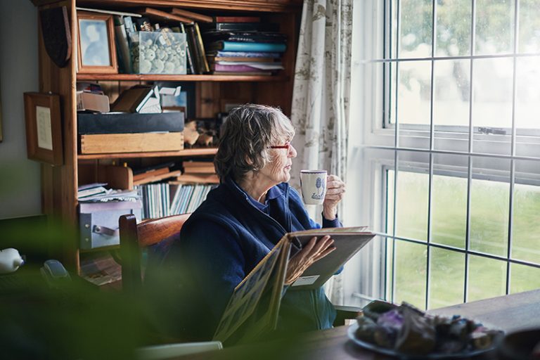 Seniorin, die aus einem Fenster hinausschaut, mit einer Tasse in der Hand und einem Fotoalbum.