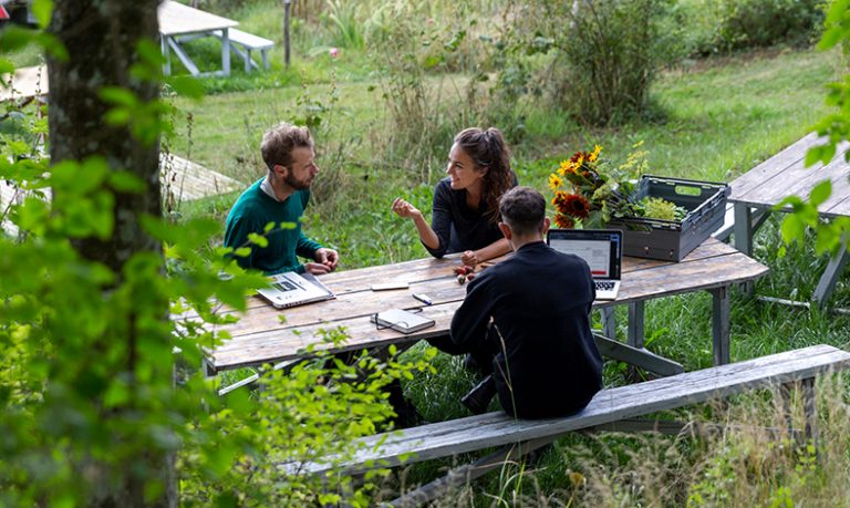 Drei junge Menschen sitzen mit Laptops in einem Garten an einem Tisch.